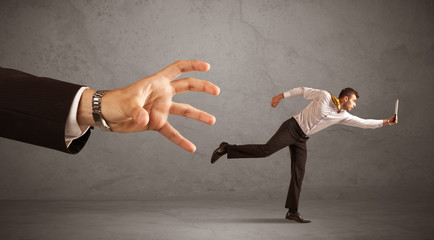Young miniature businessman running from a big hand with grey background