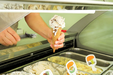 Female hand is holding a vanilla with dried fruit ice cream in waffle cone. Ice cream fridge with steel service containers in background. Worker in ice cream shop. Pastry shop. Desserts.