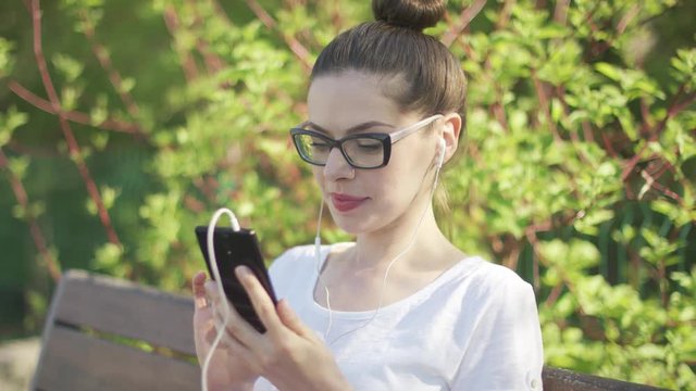 Attractive female in glasses using smartphone with headphones sitting on park bench and looking at camera