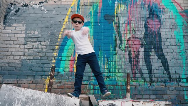 A cool 5 year old child dances in sunglasses near a brick wall with graffiti, a young street dancer,