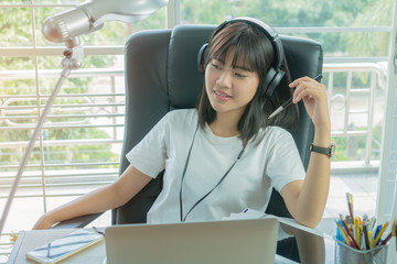 Student learning online study concept: Beautiful Asian girl sitting smile seem so happy to listening music on Headphones with her laptop on desk in home for e-learning  educational by self