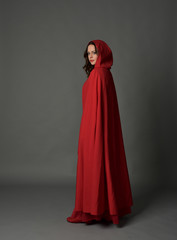 full length portrait of woman wearing red fantasy costume with cloak, standing pose on grey studio...