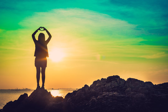 Silhouette of Heart shape being made by young woman hands with sunlight on beach. Sunset at the sea. Hand-made heart in love concept.