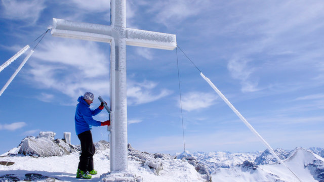 male backcountry skier on a high alpine summit preparing to write in the log book hanging on the summit cross