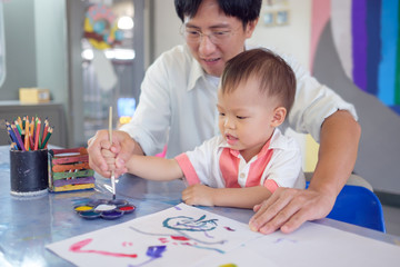 Cute smiling little Asian 18 months / 1 year old toddler baby boy child painting with brush and watercolors, Businessman father painting with son after working time, Creative play for toddlers concept