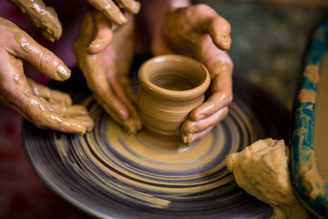 Close-up hands of potter in apron making vase from clay, selective focus. Making it together. Top view of potter teaching child to make ceramic pot on pottery wheel