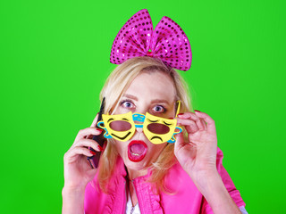 Crazy comedian blonde female in a pink suit in a funny carnival glasses is calling by phone on a colored background