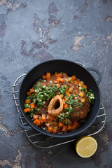 Ossobuco served with gremolata in a cast-iron pan, high angle view on a brown stone background, vertical shot with copy space