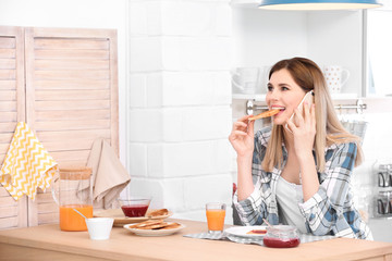 Beautiful woman eating tasty toasted bread with jam while talking on mobile phone at table