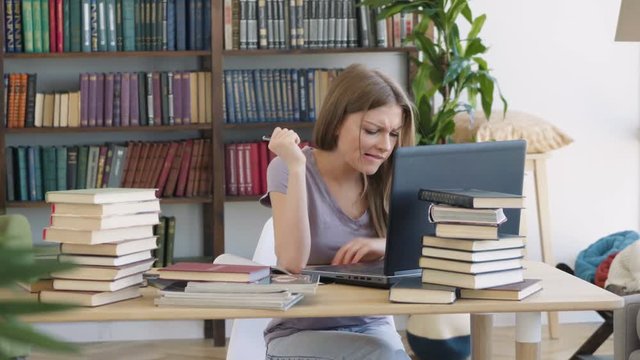 Attractive beautiful young Caucasian girl gets angry disappointed confused with studying and working on laptop in an airy light spacious library room with bookcase and bureau