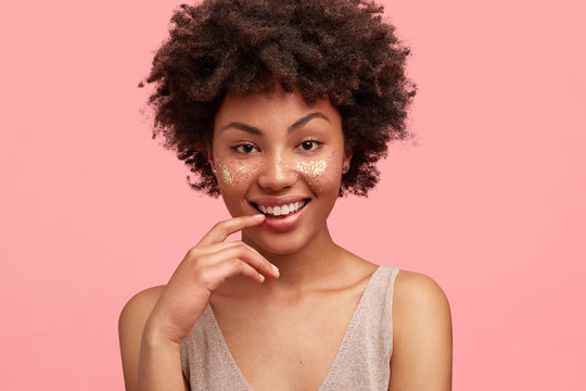 Positive African American female decorated with glitters on face, demonstrates her natural beauty, smiles joyfully, has crisp dark hair, isolated on pink background. Dark skinned female with sparkles