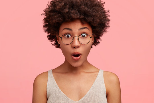 Headshot of stunned amazed beautiful African American female hears puzzled news, stares through round stylish glasses and casual t shirt, isolated over pink background. Facial expressions, ethnicity