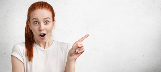 Amazed red haired young female wears casual clothing, looks with bated breath and shock, advertises...