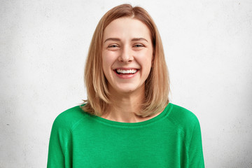 Photo of pleasant looking Caucasian woman with bobbed hairstyle, has broad charming smile, wears bright green sweater, laughs happily, hears positive funny story, isolated on white background