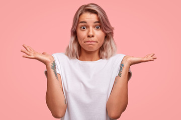 Fototapeta premium Waist up portrait of puzzled female with tattoo on arms, presses lips and shrugs hands with hesitation, wears casual white t shirt, being puzzled about something, isolated on pink background.