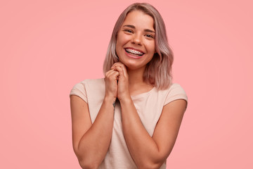 Fototapeta premium Horizontal shot of good looking female with positive smile, keeps hands together, being in good mood, rejoices winning contest, wears teeth braces, isolated over pink background. Happiness concept