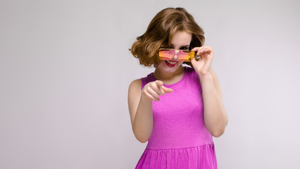 Charming young girl in pink dress on gray background. Cheerful girl in square glasses.