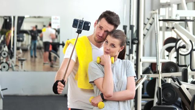 Smiling couple talking a selfie at gym. Young man and woman in sportswear taking selfie with monopod at fitness club. Woman holding dumbbell.