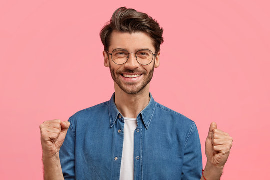 Glad male winner with cheerful expression, clenches fists in triumph, expresses his power and happiness, poses against pink background. Optimistic bearded man has happy look, gestures indoor