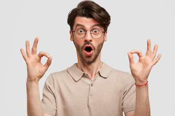 Surprised fashionable bearded guy looks with protruded eyes and widely opened mouth, shows ok sign, wears spectacles, shocked to hear something, isolated over white background. Body language.