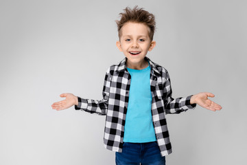 A handsome boy in a plaid shirt, blue shirt and jeans stands on a gray background. The boy spread his hands in both directions