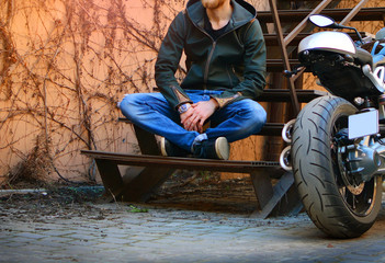 Biker life style. Man rests on the stairs near the motorcycle. Motorbike and motorcyclist on a city street yard.