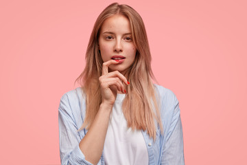 Lovely attractive female model with no make up, rests at home, thinks about how spend holidays, poses against pink studio background. European young woman looks thoughtfully and mysteriously
