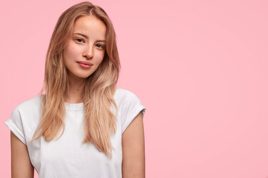 Horizontal portrait of attractive female with pleasant appearance, wears white casual t shirt, poses against pink background with copy space. Gorgeous satisfied woman student rejoies success