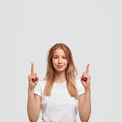 Vertical shot of Caucasian beautiful female has attractive look, indicates upwards with fore...