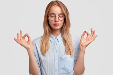 Photo of relaxed female student keeps hands in mudra sign, eyes shut, wears casual striped shirt, tries to concentrate after making difficult task or preparing diploma paper. Meditation concept