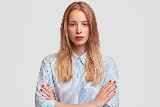 Attractive female college student keeps arms folded, wears fashionable striped shirt, looks attentively at camera, isolated over white background. Young Caucasian hipster girl with serious expression