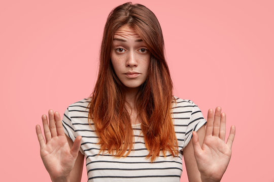 Attractive young cute woman with straight dyed hair, has freckled skin, keeps palms in foreground, rejects to do something, demonstrates her prohibition or refusal, isolated on pink background