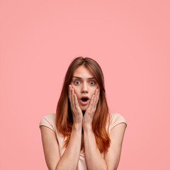 Vertical shot of surprised Caucasian female keeps mouth wide opened, looks in terror directly into camera, being scared to see something, reacts actively, isolated over pink background with copy space