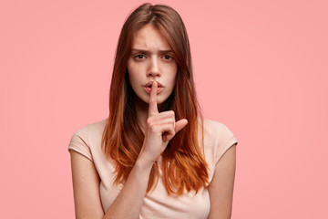 Serious beautiful female with displeased expression, makes silence sign, keeps fore finger on lips, wears casual clothes, isolated on pink background. Caucasian woman tells very private information