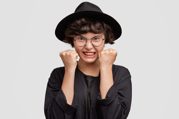 Irritated beautiful mixed race female shop assistant wears fashionable black hat, clenches fists, being annoyed with boring customers, expresses negativity and annoyance, uses gestures, isolated