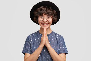 Indoor shot of pleasant looking female with glad expression, keeps hands together, stands in praying gesture, hopes for good luck, wears elegant hat and blouse, stands alone against white wall