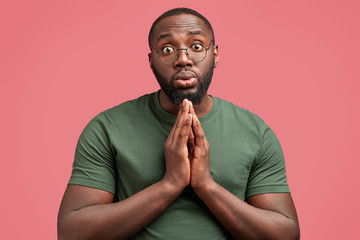 Hopeless attractive African America male stands against pink background with pleading expression, asks for forgiveness or help, wears casual t shirt. Guilty dark skinned man needs your support