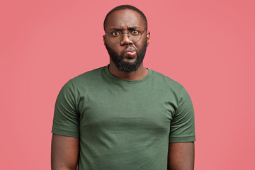 Waist up portrait of attractive dark skinned African male model curves lips and looks with discontent expression, wears glasses, tries to create new business plan, isolated on pink background