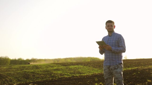 Portrait of a beautiful young man farmer student working in the field with a tractor working in a tablet, happy, dirt in a shirt, the tractor plows a soil ground field. Concept ecology. Smart farming