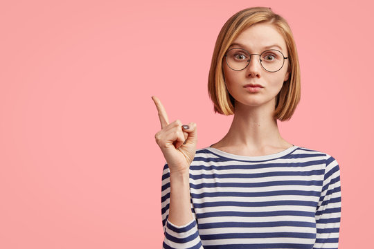 Portrait of serious stylish girl raises finger up, says: Attention please! Young stylish female teacher tries to explain something to pupils, has serious confident look, isolated on pink background