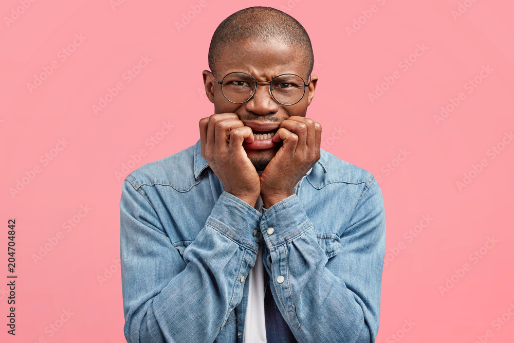 Wall mural Scraed black African American male bites finger nails nervously, being scared of something, poses against pink studio background, expresses anxiety and fear. Frightened dark skinned guy in depression