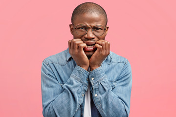 Scraed black African American male bites finger nails nervously, being scared of something, poses against pink studio background, expresses anxiety and fear. Frightened dark skinned guy in depression
