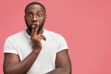 Attractive stunned thoughtful African American guy in casual clothes, has surprised pensive expression, stands against pink background with copy space for your advertising content. Ethnicity concept