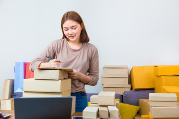 Young asian online seller businesswoman working from home writing down her customer names on her notepad and surrounded by boxes of her products,