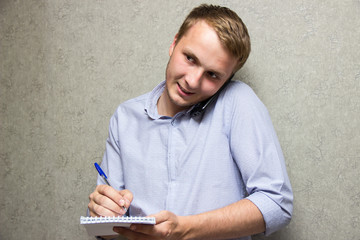 young guy in shirt talking on mobile phone with notepad and pen in hands