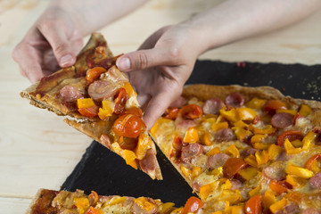 piece of pizza in hands under table with stone dish  with colored slices of vegetables close-up