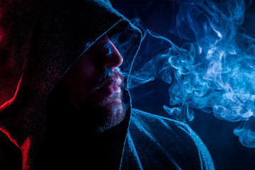 A handsome man in a black hood smokes and exhales large clouds of colored  blue and red smoke on a...