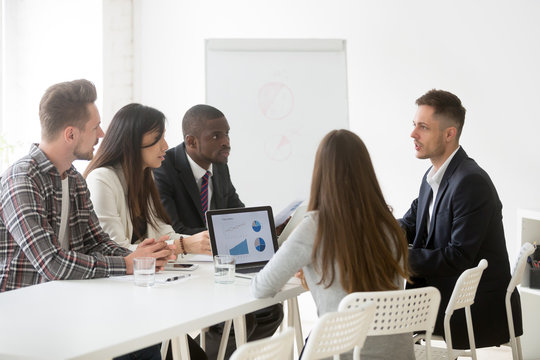 Serious multi-ethnic team listen to leader speaking at meeting, focused ceo boss coach talking to diverse surprised employees explaining solving business problem during group briefing negotiations