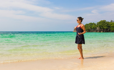 Girl on blue dress listening to music on the shore facing the sea while holding a mobile connected to earphones in the island of Koh Pha Ngan, Thailand