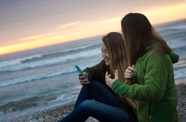 Two girls having fun with cell phone by shore with beautiful sunset behind them in La Serena, Chile. Tilt horizon